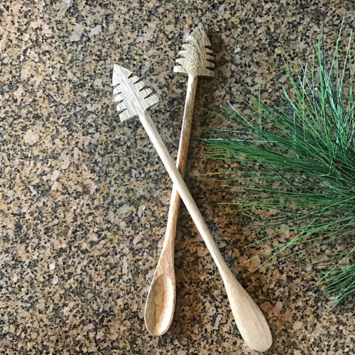 Two mango wood cocktail spoons with carved holiday tree ends, displayed crossed on a speckled granite surface beside a small sprig of pine greenery. 