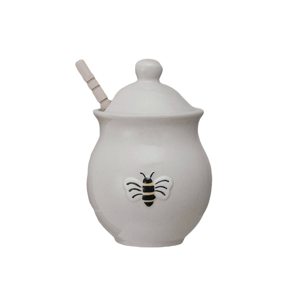 A white ceramic honey jar with a bee design, a lid, and a wooden dipper, on a wooden background.
