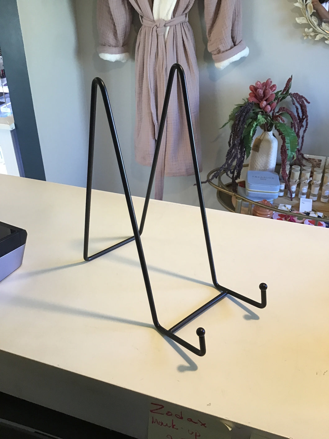 A black metal plate stand displayed on a countertop with a  robe hanging on the wall and a small table with a  floral arrangement and knick knacks.