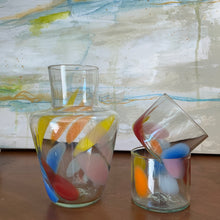 Load image into Gallery viewer, Tutti Frutti Decanter hand-blown glass with multicolor abstract design including yellow, orange, blue, red, and pink, displayed beside two matching glasses stacked on one another on wooden surface against abstract background. 