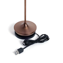 Load image into Gallery viewer, Bottom portion of the brushed copper rechargeable table light resting on its  round black magnetic charging base with a USB charging cable placed beside it, displayed on a white background. 