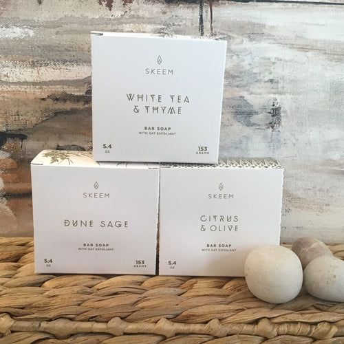 Three white boxes with gold lettering  stacked in a pyramid on a wicker platform. Small white pebbles to the side and an abstract background. 