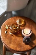 Load image into Gallery viewer, Over -head shot of a wooden table with a burning candle in a brown glass with its lid beside it, an empty pumpkin colored candle jar with trinkets inside, and its lid beside it. Out of focus grey background.  