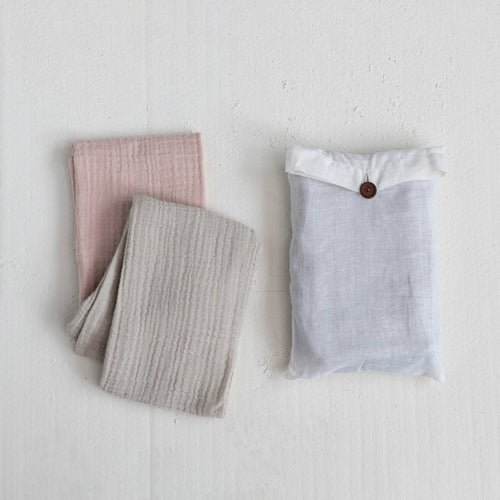 Two folded tea towels in beige and pink colors and a white bag with a brown button enclosure, placed on a white surface.