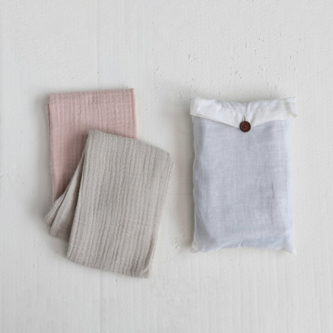 Two folded tea towels in beige and pink colors and a white bag with a brown button enclosure, placed on a white surface.
