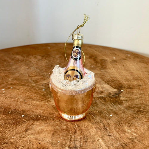 A glass replica ornament of a gold ice bucket , white ice, and a gold bottle of pink champagne. Tied gold string attached to the top of the bottle for hanging. On a brown wooden surface , against a white background. 