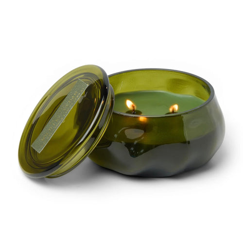 Green lit candle in a green glass container. Candle's lid is propped upon candle. Against a white background.