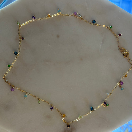 Gold chain necklace with multi-colored micro-bead charms evenly spaced around the length of the necklace. White marble background. 