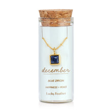 Load image into Gallery viewer, December birthstone necklace featuring a square deep blue zircon glass crystal pendant framed in 14K gold-dipped metal, displayed inside a clear glass bottle with a cork lid and a card reading "December - Blue -Zircon - Happiness + Peace," displayed against a white background. 