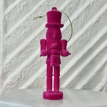 Load image into Gallery viewer, Magenta nutcracker ornament holding a small drum and drumsticks, standing upright with a gold hanging cord against a white textured background. 