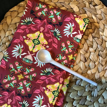 Load image into Gallery viewer, Cotton kitchen towel in burgundy with a floral pattern in yellow, pink, green, and white, displayed on a brown woven surface with a white long handle spoon on top. 