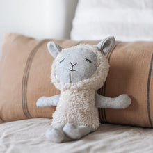 Load image into Gallery viewer, Gray-and-cream fabric sheep doll with a fluffy textured body and black embroidered sleepy eyes, displayed sitting against a tan pillow with black stripes. 