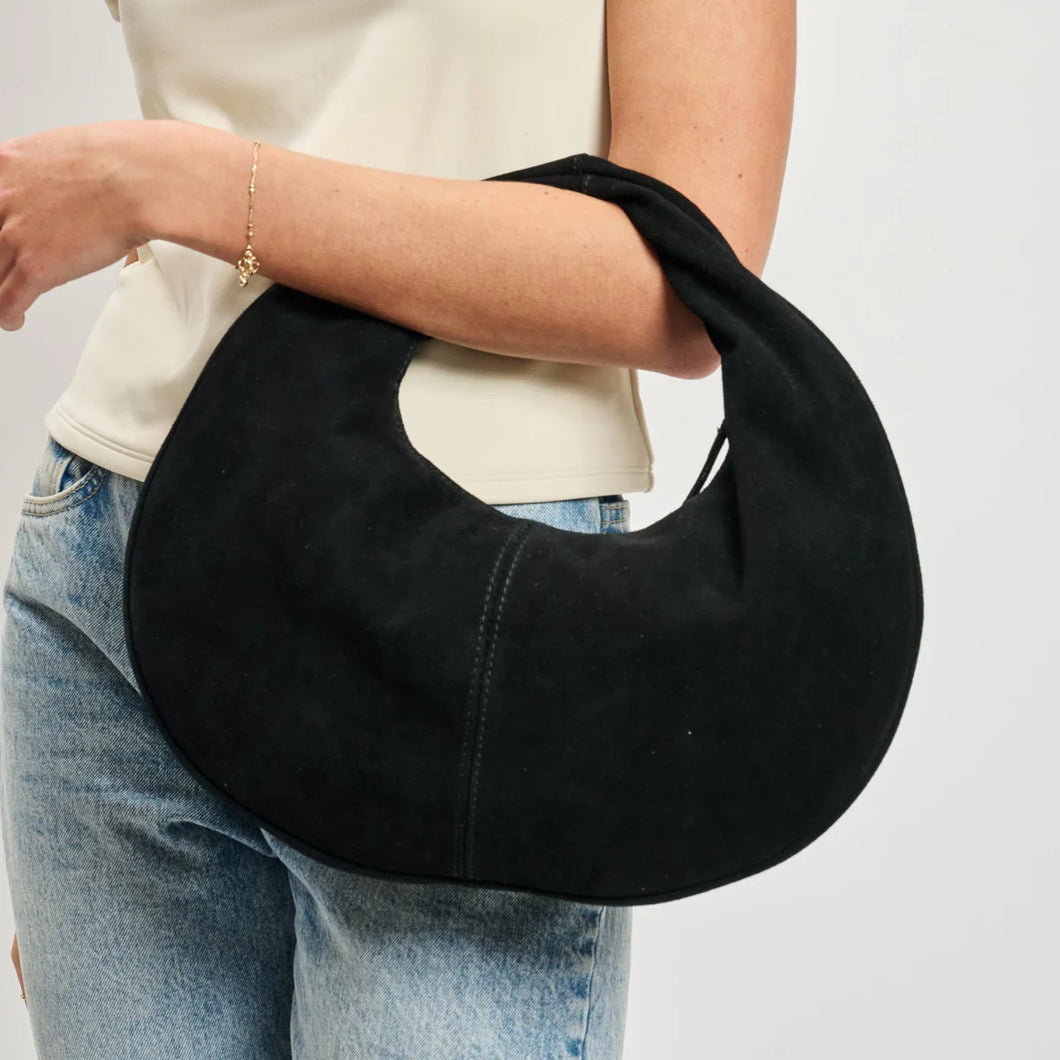 Black suede crescent-shaped hobo bag held on model's forearm, paired with a white top and light-wash jeans against a white background. 