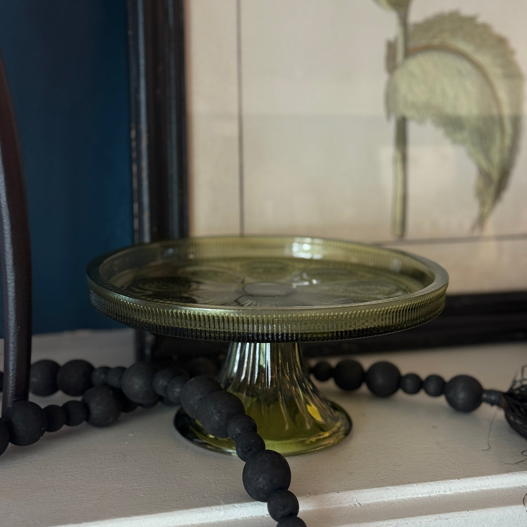 Olive colored glass cake stand with a ribbed lip that is partially black. Cake stand is sitting on a white surface with a black bead garland and a blurred framed picture in the background, against a dark blue background. 