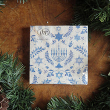 Load image into Gallery viewer, IHR cocktail-sized paper napkin sets with a Hanukkah design featuring blue menorahs, Stars of David, dreidels, and leafy motifs on a white background. Displayed on a wooden surface surrounded by green garland. 