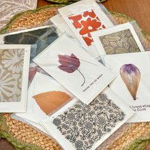 Load image into Gallery viewer, Colby Cards, lifestyle image of overlapping greeting cards on white Lokta paper featuring real pressed flowers alongside printed botanical designs, including a central “Thank You So Much” card, displayed in clear plastic sleeves on a brown woven mat with green scalloped edging atop a wooden table.