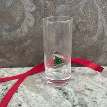 Load image into Gallery viewer, ZODAX Holiday Shot Glasses, lifestyle image of a tall clear shot glass with a thick base and a raised green Christmas tree motif with red accents, resting on a gray marble surface with a red ribbon and a patterned gray background.