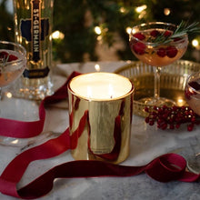 Load image into Gallery viewer, Lit metallic gold candle on a white marble surface with a long red ribbon, a gold tray with a cocktail glass filled with a light colored beverage and cranberries, a bottle of St. Germain, and a blurred lit Christmas tree in the background.  