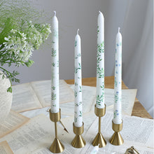 Load image into Gallery viewer, Four white taper candles with botanical and floral designs in gold candle holders, displayed on scattered paper pages with a white floral arrangement in the background. 