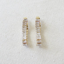 Load image into Gallery viewer, Pair of gold IP-plated earrings with curved rows of clear baguette-cut CZ stones displayed on a soft white textured background. 