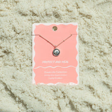 Load image into Gallery viewer, Sterling silver-dipped necklace featuring a small scallop shell charm pendant and a chain with delicate beaded accents, displayed on a light pink &quot;Protect and Heal&quot; Ocean Life Collection card by Lucky Feather with a white wavy border, shown resting on white textured sand. 