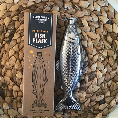 Silver flask in the shape of a fish next to it's brown cardboard box that has the flask printed on it. Sits on a brown braided placemat. 