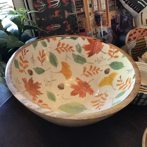Wooden bowl with a white enamel interior featuring a harvest design with green, yellow, and orange leaves, displayed on a black surface. In the background are a green plant, an opened book, a stack of blue-and-white checkered bowls, and an orange-and-brown pillow. 