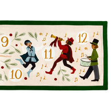 Load image into Gallery viewer, Close-up of the gold &quot;10&quot;, &quot;11&quot;, and &quot;12&quot; illustrations from the 12 Days of Christmas table runner featuring a leaping lord, a piper playing a horn, and a drummer with a marching drum, surrounded by scattered green foliage on a white background with a green border. 