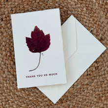 Load image into Gallery viewer, Colby Cards, lifestyle image of a white Lokta paper greeting card featuring a real pressed burgundy leaf above “Thank You So Much” in dark burgundy lettering, displayed with a white envelope on a brown woven mat.
