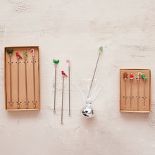 Load image into Gallery viewer, Two sets of holiday glass drink stirrers with festive toppers including holly leaves,  red cardinals, Christmas trees, candy canes, a Santa hat, and a gingerbread figure, displayed in brown boxes and arranged beside a martini glass on a textured cream background. 
