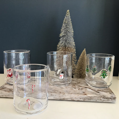 A set of four tumbler glasses each with a different holiday design : red and white candy canes, red and white Santas, white snowmen with black top hats and a red scarves, and green Christmas trees with white and red ornaments. Santa , Snowman, and Christmas tree tumblers are sitting on a cream and brown marble slab. Candy cane tumbler is sitting on a white surface. Two decorative frosted Christmas trees against a blue background. 