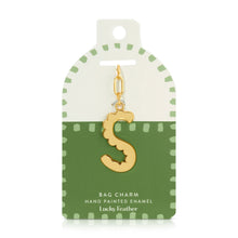 Load image into Gallery viewer, 14K gold-plated hand-painted enamel initial bag charm featuring the letter S with a scalloped edge design and a brushed gold finish, attached to a key ring and clasp. Displayed on a white and green Lucky Feather card with light green dotted brush-stroke borders labeled &quot;Bag Charm Hand Painted Enamel.&quot; Displayed against a white background. 