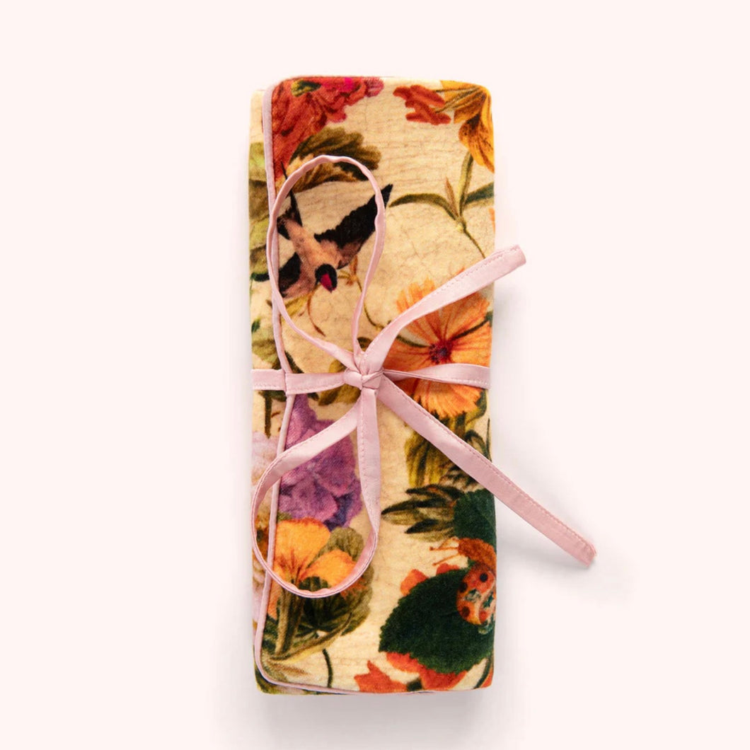Velvet jewelry roll in a cream floral pattern featuring colorful blooms and a bird motif, tied with a pink ribbon and displayed against a white background. 