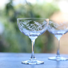 Load image into Gallery viewer, Stemmed wine glass with etched leaves on a wooden platform. Another wine glass is behind it blurred in the background. Background is a a green nature scene. 