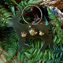 Load image into Gallery viewer, Hunter green deer shaped metal ornament with three gold jingle bells. A silver metal ring attached to the deer for hanging. Ornament is lying on a bed of green ferns , and a charred log. Brown woven basket partially visible in top right corner. 