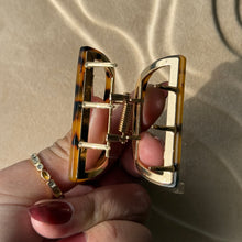 Load image into Gallery viewer, Tortoiseshell half-moon metal claw clip with gold hardware held in a hand against a beige textured surface with soft curved light reflections. 
