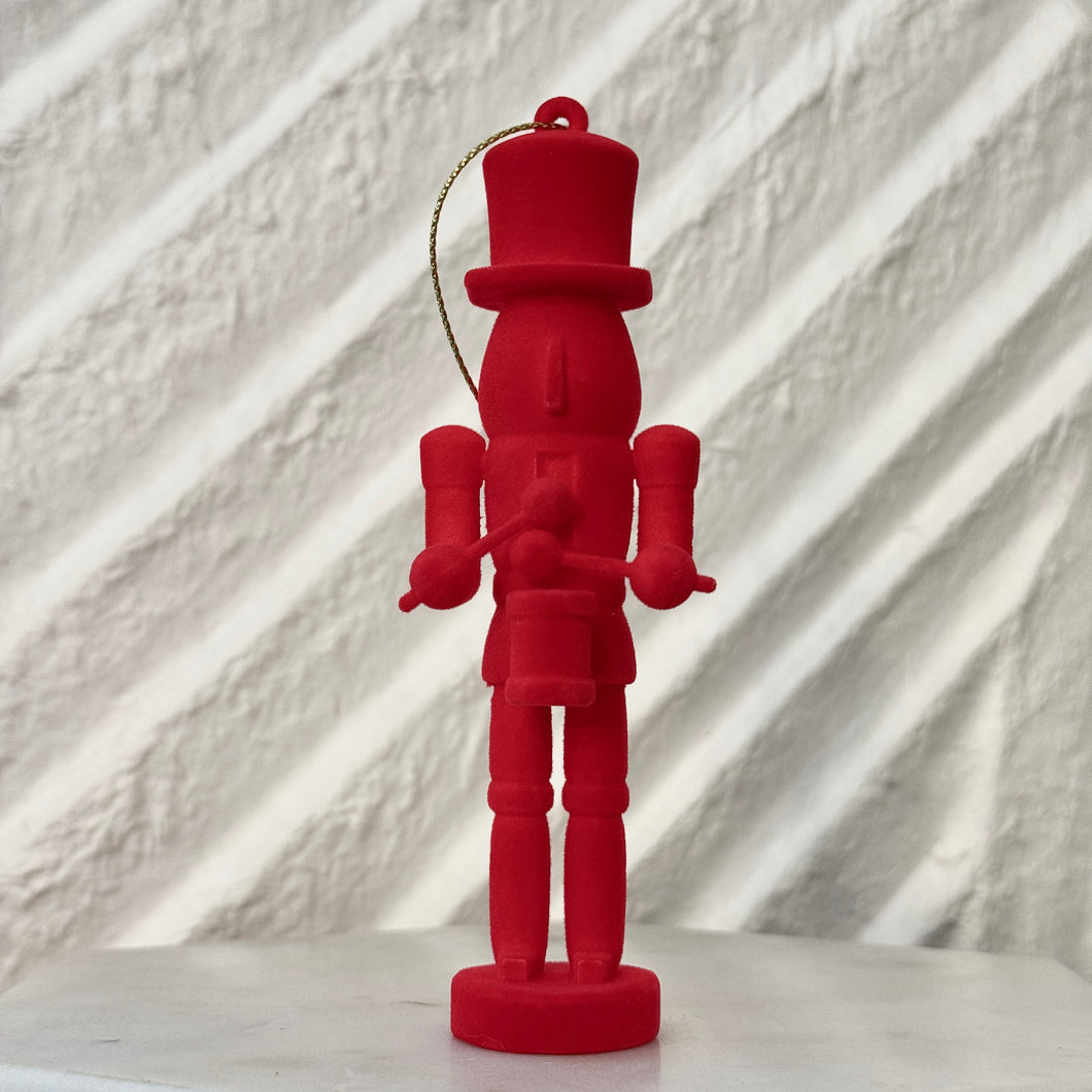 Red nutcracker ornament holding a small drum and drumsticks, standing upright with a gold hanging cord against a white background. 