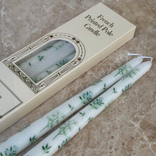 Load image into Gallery viewer, Two white taper candles with green-and-blue botanical designs on a brown marble surface, next to their packaging.