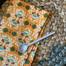 Load image into Gallery viewer, Cotton kitchen towel in orange with a floral pattern in green, cream, and burgundy, displayed on a brown woven surface with a white long handle spoon resting on top.