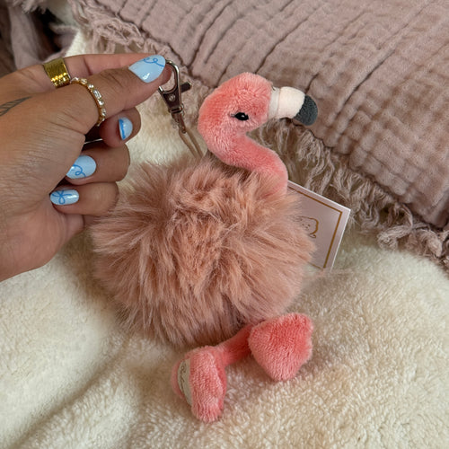 Person with blue manicured nails holding a pink plush flamingo with a fuzzy mauve- colored middle with a silver clasp. On a soft white surface with a pink textured textile in the background.