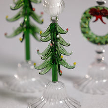Load image into Gallery viewer, Close-up of a green glass Christmas tree taper candle holder with curled layered  glass branches and red and yellow accents, with a matching tree holder and a green wreath holder with a red bow softly blurred in the background on a white surface. 