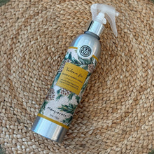 Load image into Gallery viewer, Balsam &amp; Fir, lifestyle image showing an overhead view of a silver-tone room and fabric spray bottle with a translucent white plastic trigger, a circular Mangiacotti emblem, and a beige wraparound label featuring green evergreen branches, brown pinecones, and a gold-colored panel with the scent name and product type in dark lettering, arranged diagonally on a brown woven mat.