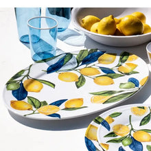 Load image into Gallery viewer, White melamine oval serving platter with a yellow lemon and green leaf pattern, displayed with matching dinnerware, a white bowl full of lemons, and blue tumbler glasses, displayed on a white background.  