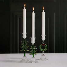 Load image into Gallery viewer, Three glass taper candle holders shaped like green Christmas trees and a wreath, each holding a lit white taper candle, displayed on a white surface against a softly blurred dark background. 