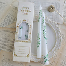 Load image into Gallery viewer, Two white taper candles with green botanical designs displayed on an ornate white plate over scattered paper pages, next to their packaging. 