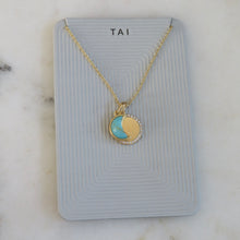 Load image into Gallery viewer, Gold-plated necklace with a turquoise-colored crescent moon charm on top of a round gold pendant with a crystal accent border, displayed on a light gray jewelry card with "TAI" in black print on a white marble surface. 