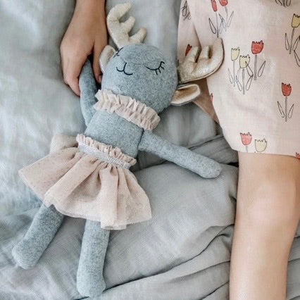 Gray wool-blend deer doll with sparkly gold antlers and ears, black embroidered sleepy eyes, a beige ruffle collar,  and a matching tulle skirt, displayed in a child's hand on a gray blanket. 