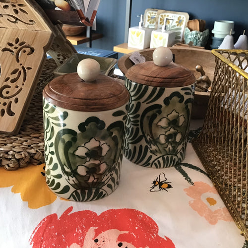 Two black and white floral canisters with wooden lids on a table with various objects in the background. 