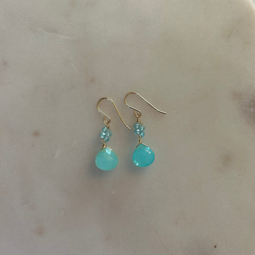 A pair of gold drop earrings with four blue beads and a blue teardrop gem. White marble background. 