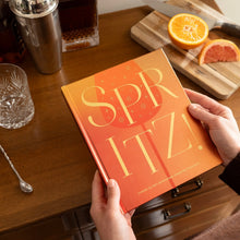 Load image into Gallery viewer, "Spritz!" drink recipe book with bold gold text on orange background shown near citrus and bar tools.